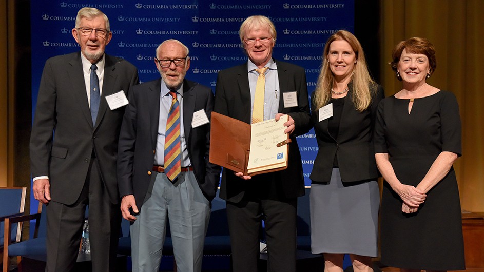 3 men and 2 women with middle man with blonde hair, blue shirt, suite jacket yellow tie holding signed document