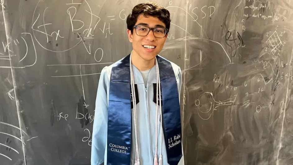 Luca Nashabeh wearing Columbia University regalia in front of a blackboard with physics equations before his Columbia College class day ceremony. 